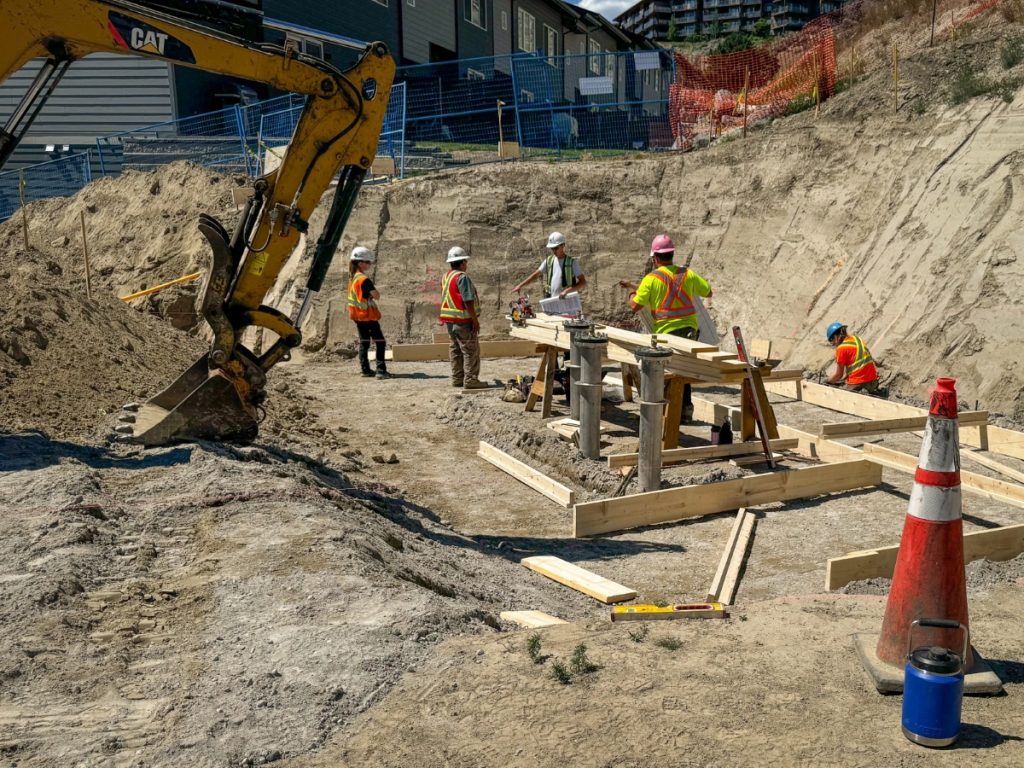 Construction crew setting foundation formwork at the Sagewood Booster Station construction site in Kamloops, BC, with a CAT excavator and Sagewood neighbourhood homes visible in the background.